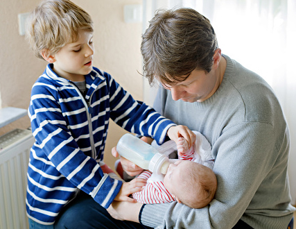 A boy helps an man bottle feed an infant